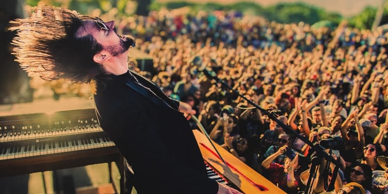 Performer at keyboard with massive crowd visible behind under warm stage lighting.