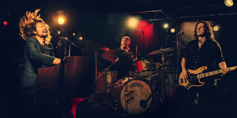 Three-piece band performing on stage under warm stage lighting, with lead singer at microphone, drummer, and bassist.