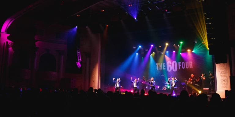 Band performing on stage under vibrant multi-coloured stage lighting with crowd silhouettes in foreground.
