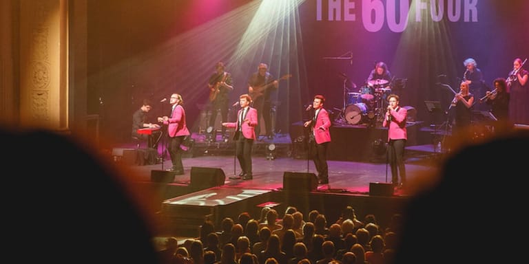 Musicians performing on stage in matching pink attire, lit by purple and gold spotlights, with audience members in foreground.