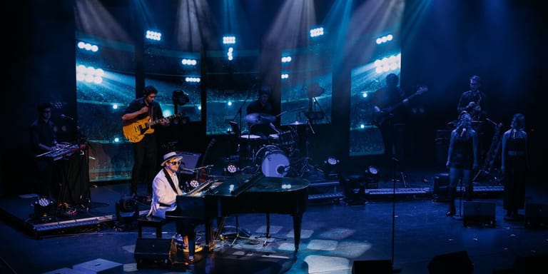 Pianist performs at a grand piano centre-stage amid a five-piece band under blue theatrical lighting.