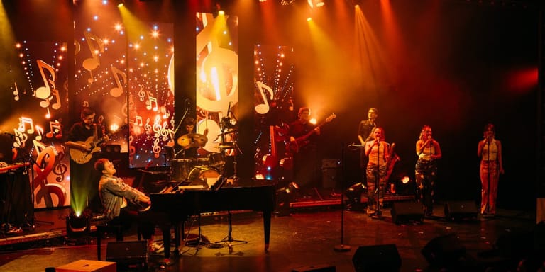 Grand piano and ensemble performers bathed in warm amber stage lighting during a theatrical production.