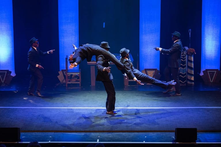 Dancer in black suit bends backwards dramatically on stage whilst band members perform under blue lighting.