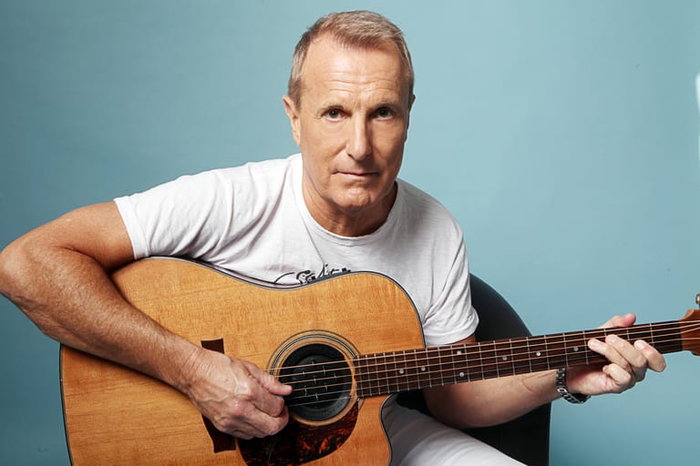 Musician holding an acoustic guitar against a blue background, wearing a white t-shirt.