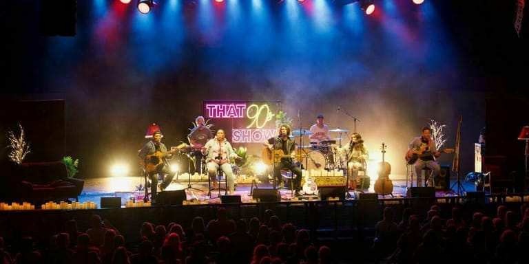A band performs on an illuminated stage before a crowd, with purple and blue stage lighting and decorative elements overhead.