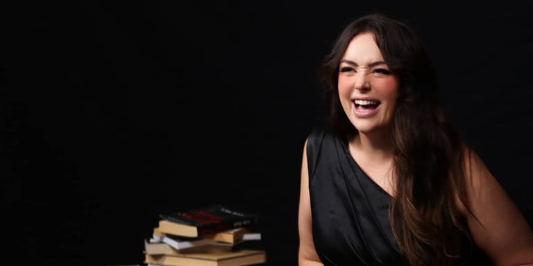 Woman with long dark hair smiling at camera beside a stack of books against a black background.