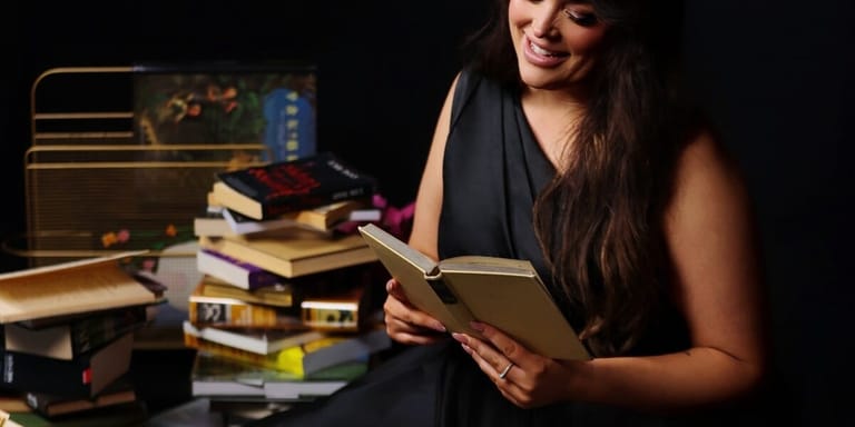 Woman with long dark hair reading an open book surrounded by stacked books and a desk lamp.