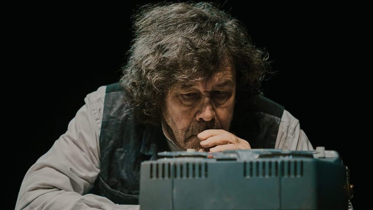 Man with curly hair and beard leaning over a grey electronic keyboard against a black background.