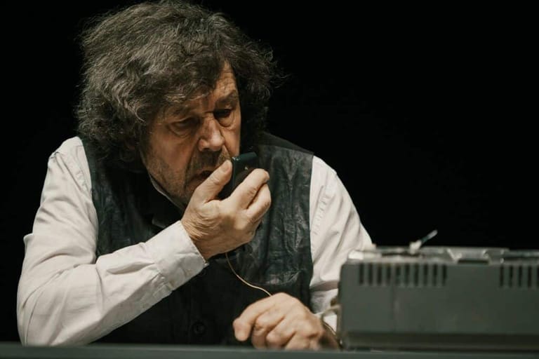Man with grey hair in white shirt and dark vest, hand to face, seated at keyboard against black background.