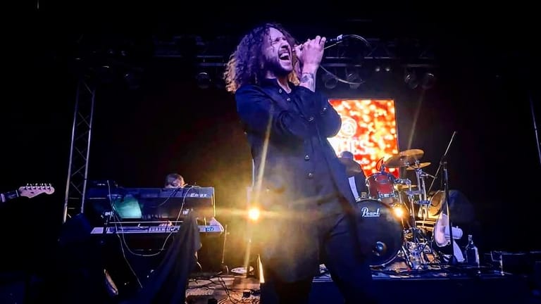 Performer with curly hair singing at microphone on stage illuminated by warm spotlights, with keyboard and drums visible.