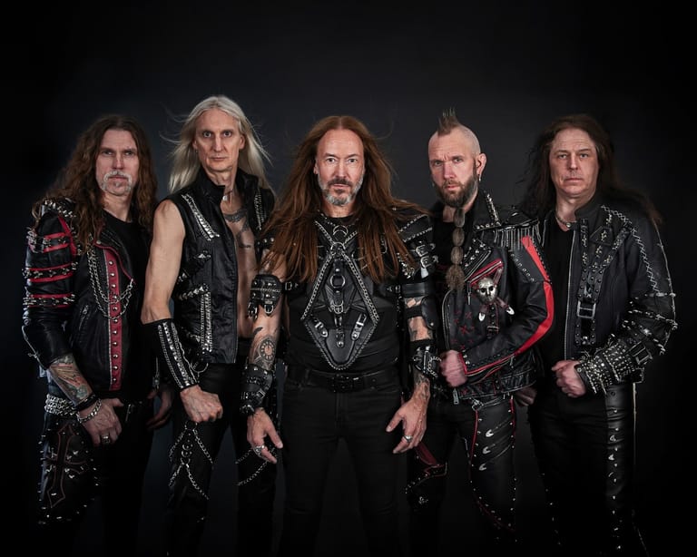 Five men in ornate black leather and studded stage costumes posed against a dark background.