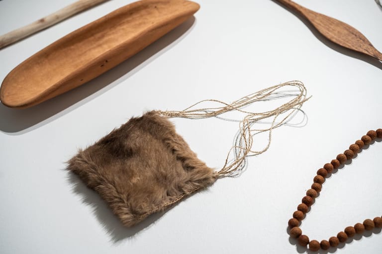 Indigenous artefacts including wooden tools, fur pouch and beaded necklace displayed on white background.
