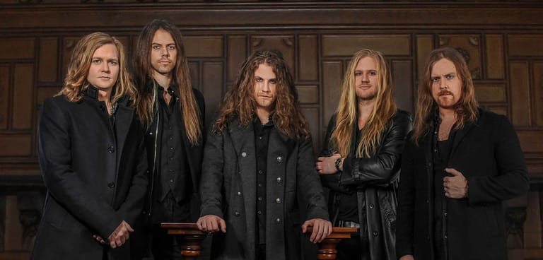 Five musicians with long hair in black clothing stand against a wooden panelled backdrop.
