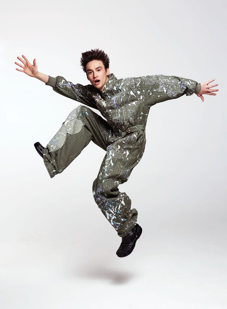Person in silver sequined suit mid-leap against white background, arms outstretched in exuberant pose.