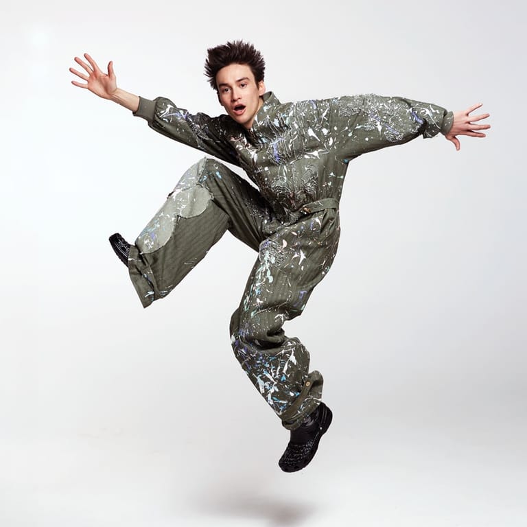 Performer in a sparkly grey suit leaps mid-air with arms outstretched against a white background.
