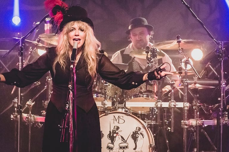 Female performer with long blonde hair and red flower accessory singing at microphone with drummer and band behind her on stage.