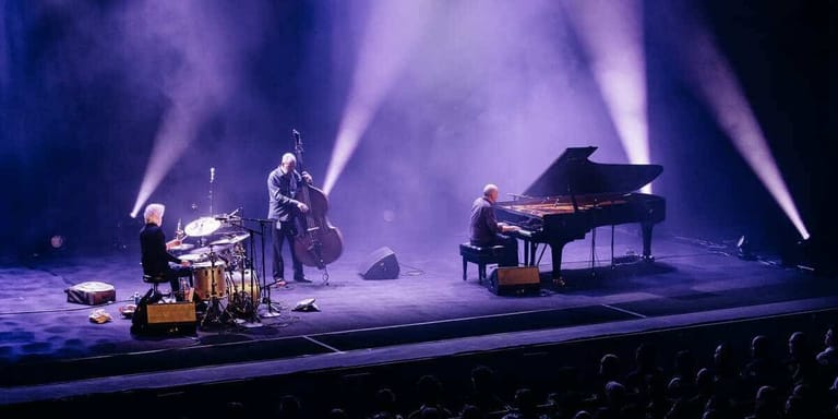 Three musicians perform on a concert stage bathed in purple lighting, with a grand piano, drums, and bass player under dramatic spotlights.