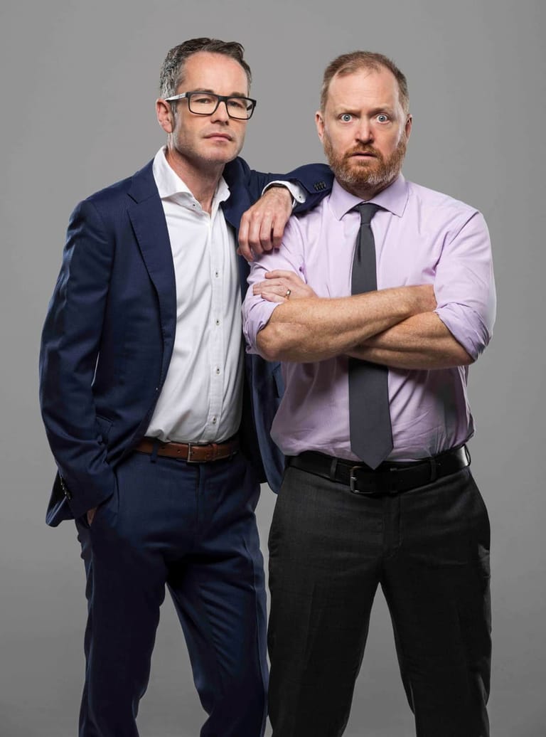 Two men in business attire pose together against a neutral grey background, one wearing a navy suit and glasses, the other in a pink shirt and dark tie.