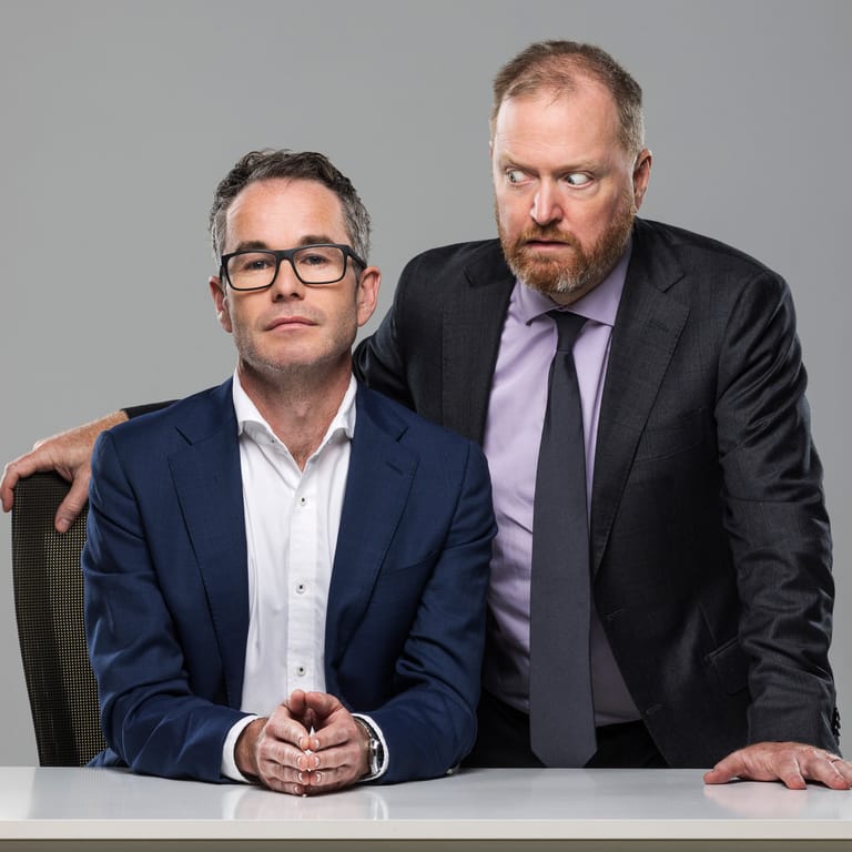 Two men in business attire pose together at a white table against a neutral grey background.