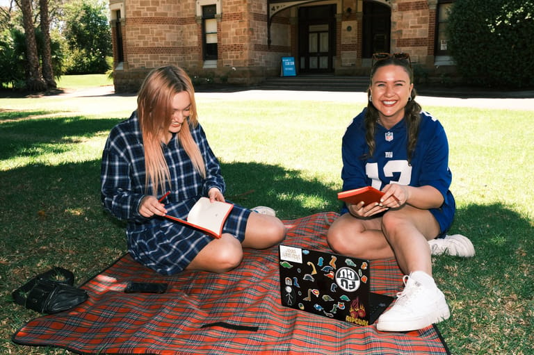 Two people sit on a picnic blanket on grass outside a brick building, sharing food from a box.