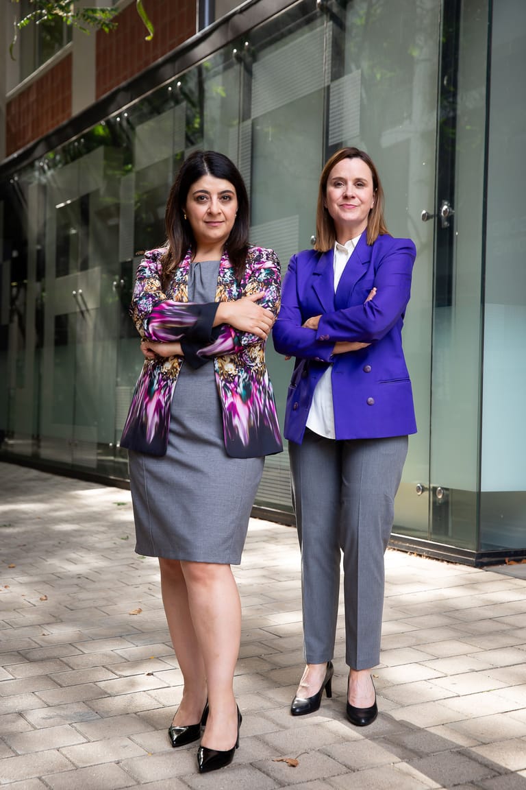 Two women stand with arms crossed outside a modern glass-fronted building, one wearing a patterned jacket and grey skirt, the other in a purple blazer and grey trousers.
