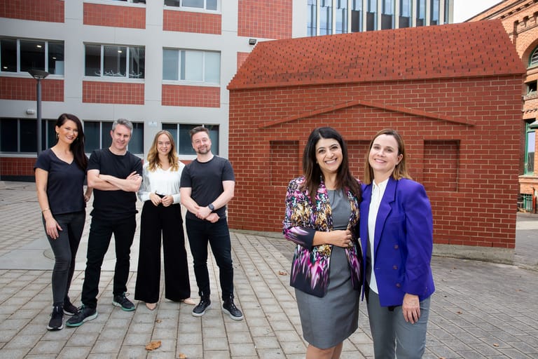 Group of eight people standing in a contemporary urban courtyard beside a red brick building.