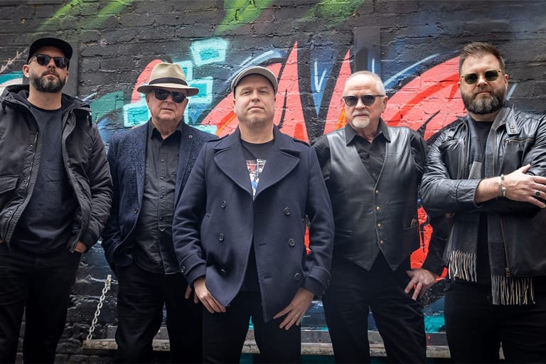 Five men in dark leather jackets stand together against a vibrant street art backdrop with neon-lit geometric shapes.