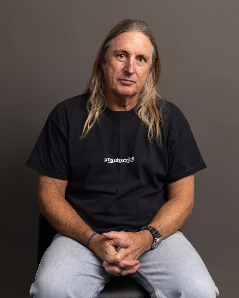 Man with long blonde hair wearing black t-shirt and light jeans sits against neutral background.