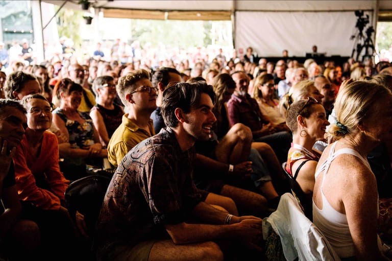 Dense crowd of festival-goers seated indoors under warm lighting, many wearing casual summer clothing.