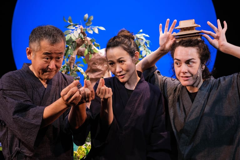 Four performers in black clothing playfully pose with props against a bright blue theatre backdrop.