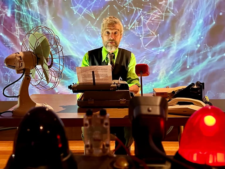 Man in suit seated at desk surrounded by vintage electrical equipment and glowing coloured projections.