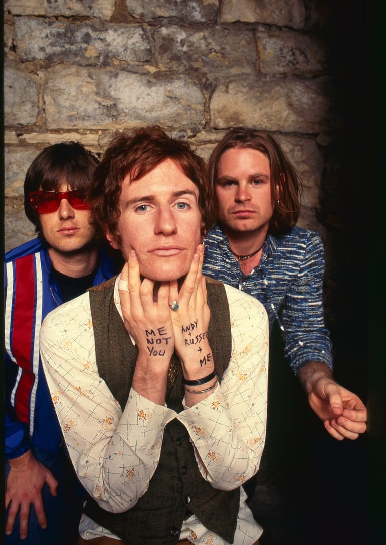 Three band members pose against a weathered brick wall, with the front member displaying text written on his hands.