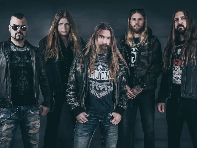 Five musicians in dark leather and metal jewellery stand against a grey backdrop in a professional band portrait.