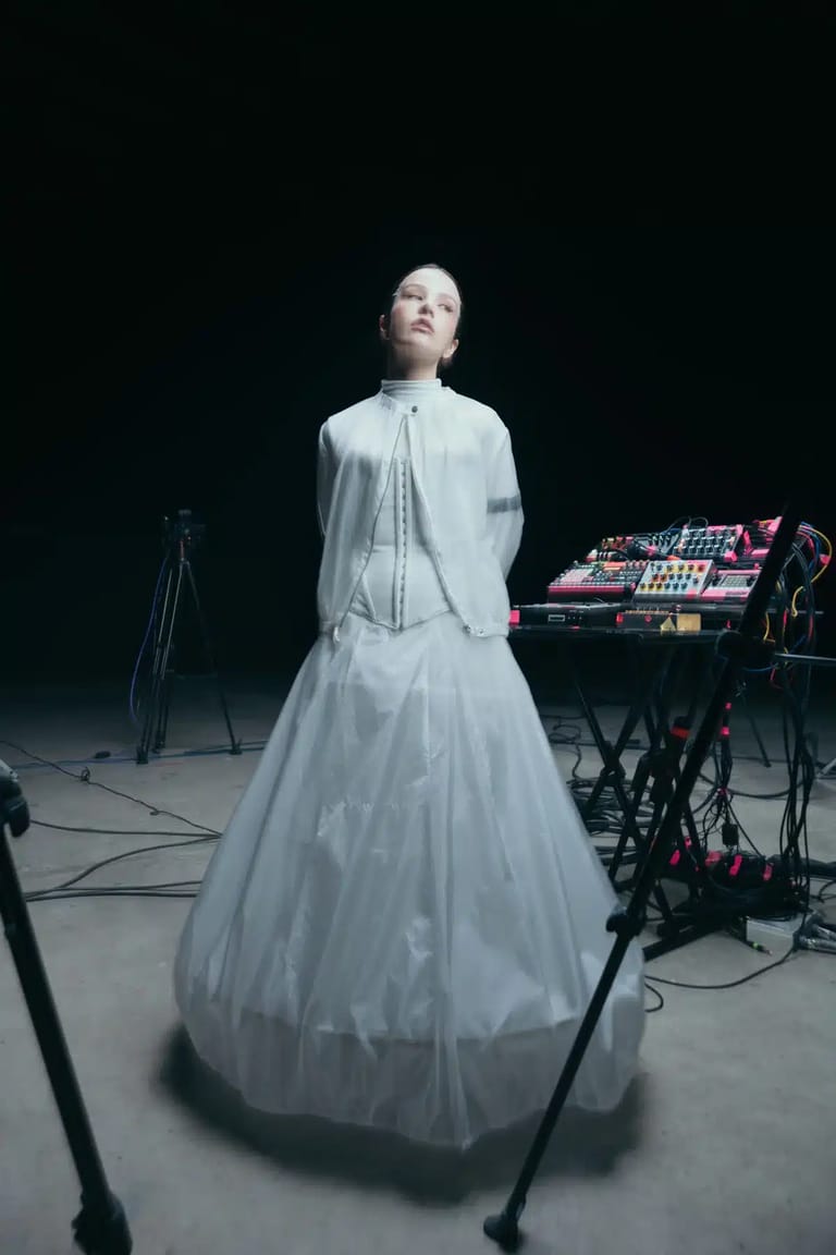 Performer in flowing white gown gazes upward beside synthesiser on dark stage.