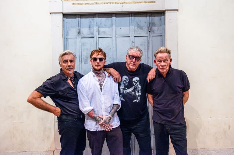 Four men in sunglasses and dark clothing stand together against a white wall with a blue door behind them.