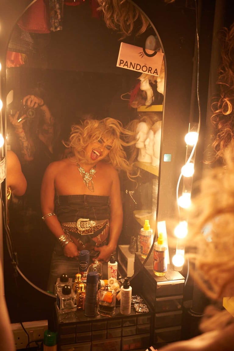 Performer in a strapless black top and jewelled belt reflected in a Hollywood-style vanity mirror with illuminated bulbs.