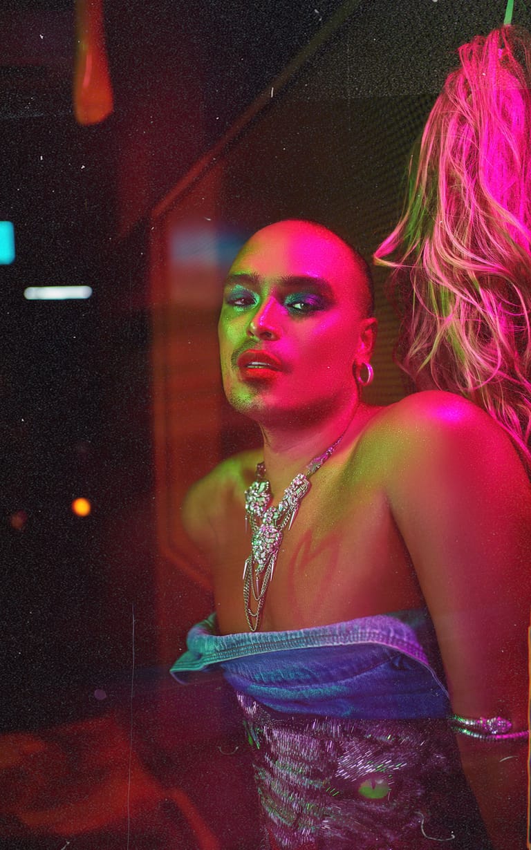 Performer in neon pink and green makeup wearing jewelled accessories under vibrant stage lighting.