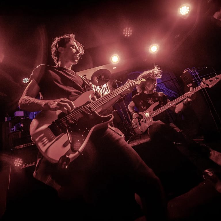 Guitarist performs under warm stage lighting with band members visible in burgundy-tinted concert setting.