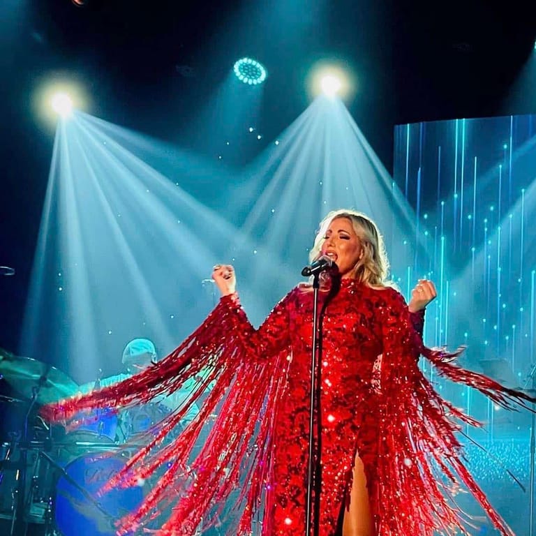 Performer in a red sequined gown with dramatic wings sings centre stage under blue and white theatrical spotlights.