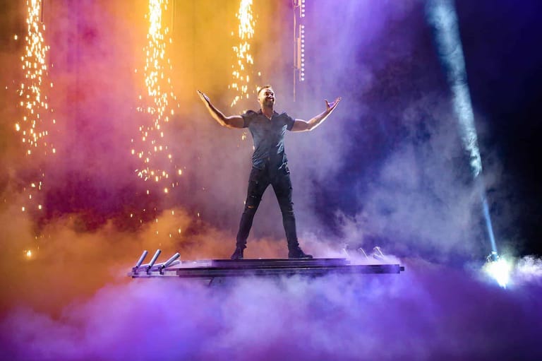 Performer stands on stage with arms raised, surrounded by cascading golden lights and vibrant purple and pink smoke effects.