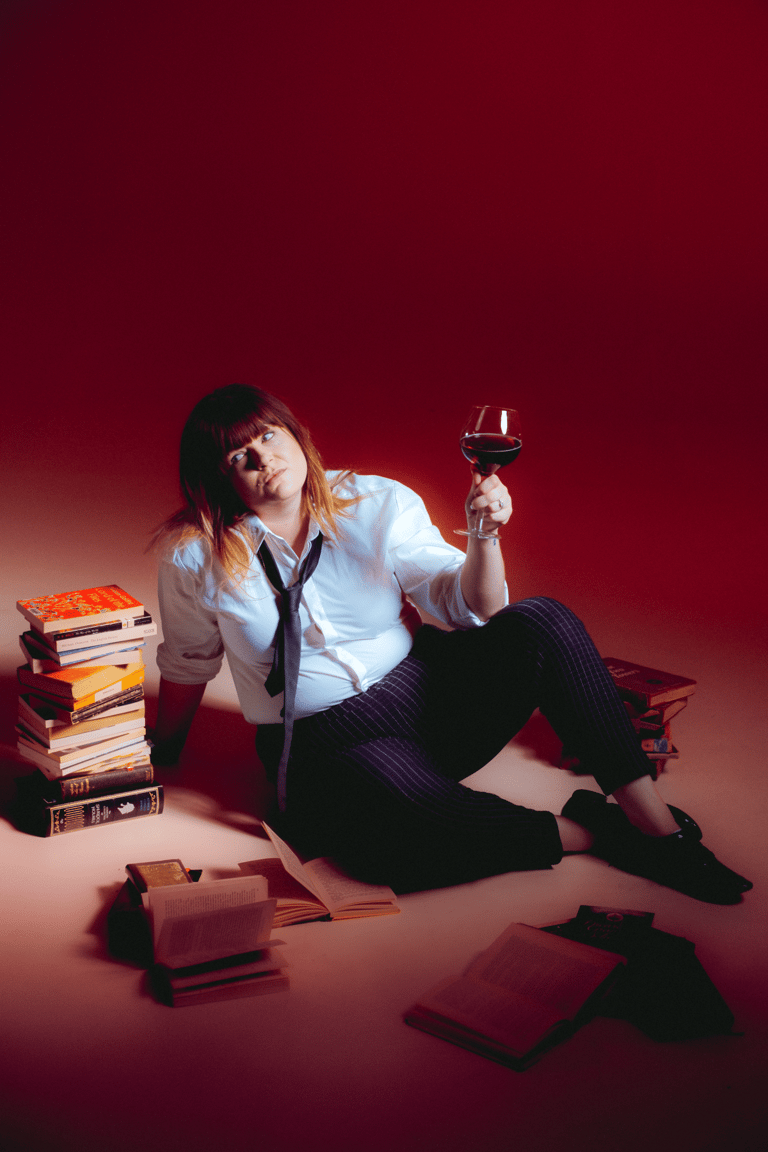 Person seated against a deep red background surrounded by stacked books, holding a wine glass with a contemplative expression.