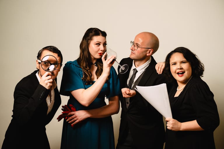 Four performers in business attire strike exaggerated comedic poses against a neutral backdrop.