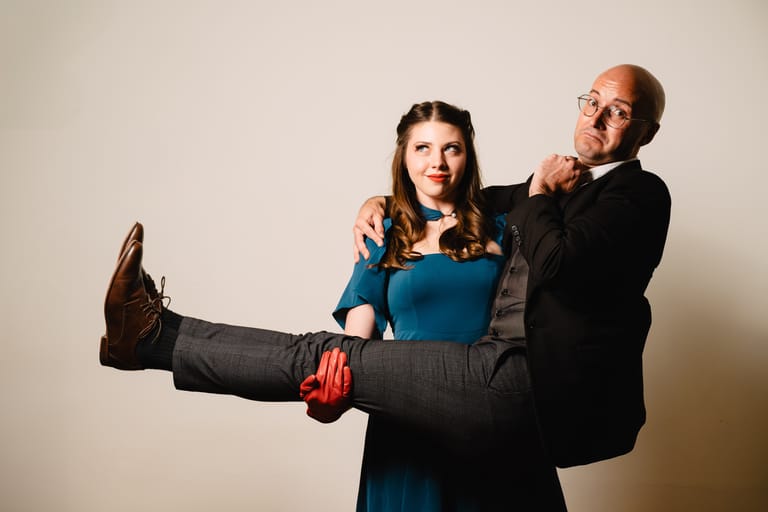 Man in black suit carries woman in blue dress with leg raised against neutral backdrop.