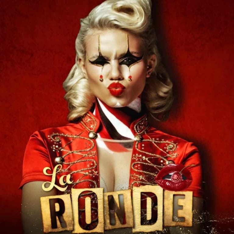 Performer in bright red ornate jacket with platinum blonde hair and dramatic makeup against red backdrop, "La Ronde" text overlay.