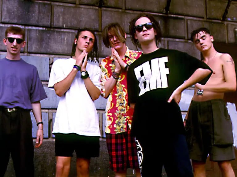 Five young people in casual 1990s-style clothing stand against an industrial brick wall, wearing sunglasses and striking posed attitudes.