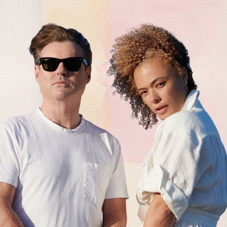 Two people in white t-shirts stand against a soft pink and yellow gradient background, one wearing dark sunglasses.
