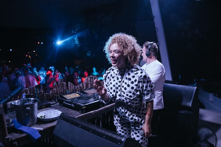 DJ in black-and-white patterned shirt performing at turntables before a crowd under blue stage lighting.