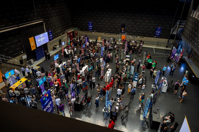 Overhead view of a crowded convention or festival floor with hundreds of attendees mingling amongst exhibition stalls and blue-lit displays.