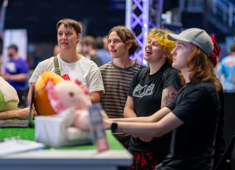 Four people in casual clothing hold a glowing ball at an indoor event with stage lighting and crowds visible behind them.