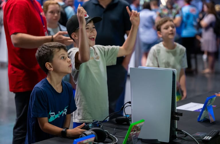 Young children celebrate at a gaming or technology event, one with arms raised in excitement beside a computer monitor.